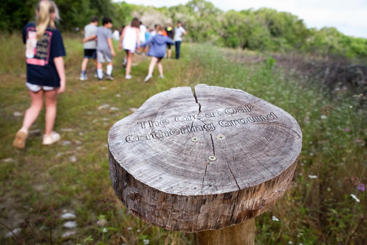 Photo of a sign made from a tree ring that says "The great oak gathering ground". Kids are seen walking on the trail in the background.
