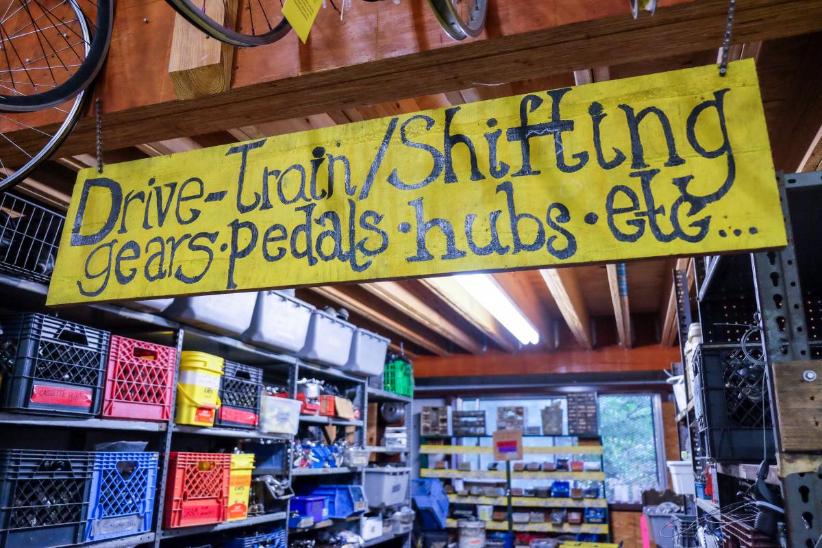 Interior of bicycle shop. Sign reads "Drive-train/Shifting gears, pedals, hubs, etc..."