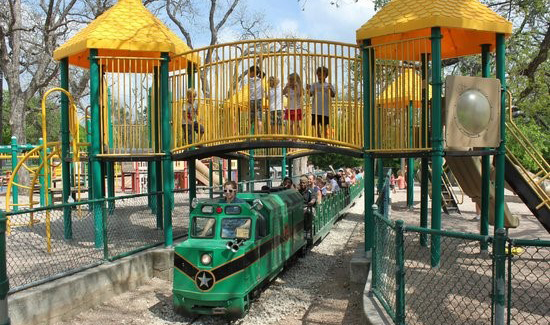 Playscape with bright yellow shade cones and bars with kids playing on the upper deck. A green train is going through the playscape.